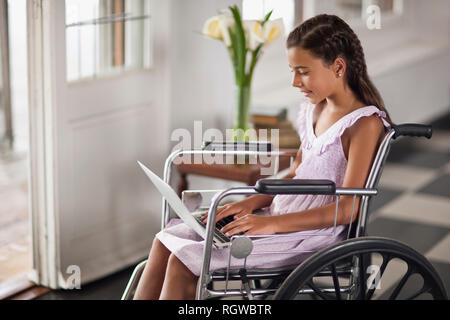 Giovane ragazza seduta in carrozzella digitando su laptop. Foto Stock