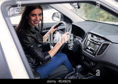 Bella sorridente driver femminile in giacca nera seduta all'interno auto e trattenendo il volante. Uno sguardo alla finestra del veicolo a felice attraente brun Foto Stock