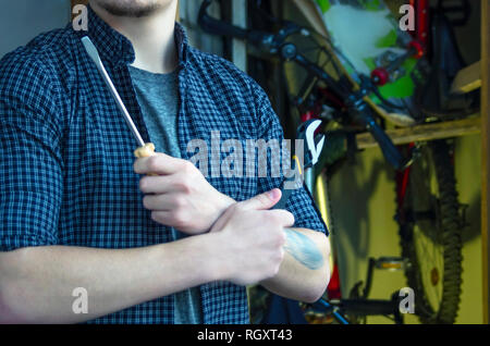 Barbuto di Classe Giovane con un tatuaggio in una T-Shirt e plaid Shirt si prepara alla Manutenzione bicicletta. Con le braccia incrociate azienda strumenti. Biciclette e pattini in Foto Stock