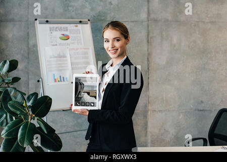 Attraente sorridente imprenditrice azienda digitale compressa con biglietti sullo schermo Foto Stock