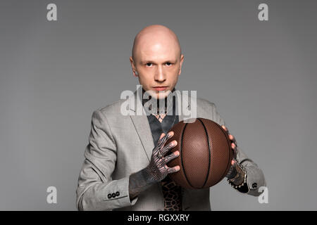 Grave bald tatuato uomo in tuta holding basket ball e guardando la telecamera isolato su grigio Foto Stock