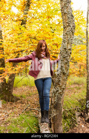 Una giovane ragazza con i capelli rossicci saldi su un tronco caduto a terra in una foresta autunnale Foto Stock