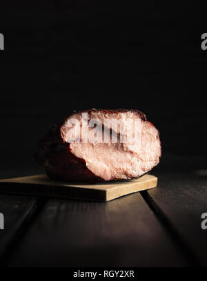 Close up foto scuro illuminato di prosciutto affumicato su tavola di legno con sfondo nero. Fresca a fette di carne affumicata su un tavolo di legno (isolato shot). Foto Stock