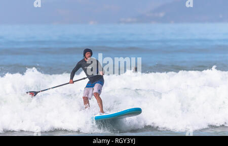 Paddleboarding su un SUP o Stand Up Paddle ski cavalcare le onde verso la spiaggia su una soleggiata giornata autunnale a Muizenberg di Città del Capo in Sud Africa Foto Stock