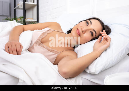Ha risvegliato la bella giovane donna in elegante nightie giacente sulla biancheria da letto bianca e sorridente Foto Stock