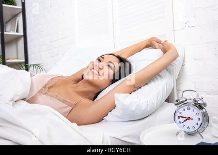 Ha risvegliato la donna sorridente in eleganti nightie giacente sulla biancheria da letto bianca e stretching Foto Stock