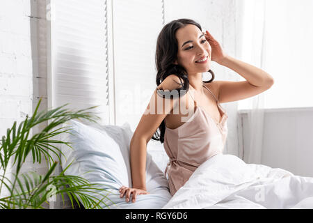 Bella giovane donna asiatica in eleganti nightie seduta a letto e di toccare i capelli con la mano Foto Stock