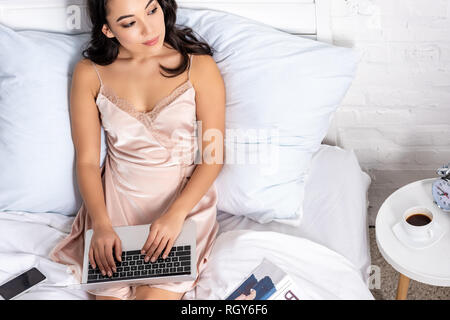 Giovani asiatici freelancer in eleganti hightie utilizzando computer portatile mentre stare a letto e guardando alla finestra Foto Stock