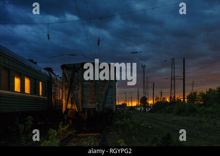 Tramonto colorato con cielo cupo presso la stazione ferroviaria in estate. Le finestre di un'autovettura sono accese. Foto Stock