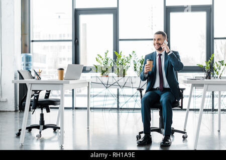 Allegro imprenditore tenendo bicchiere di carta mentre si parla sullo smartphone in un ufficio moderno Foto Stock