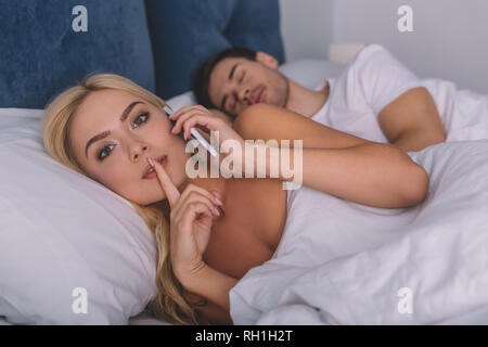 Giovane e bella donna che parla tramite smartphone e guardando la telecamera con il dito sulle labbra mentre il marito dorme nel letto, concetto segreta Foto Stock