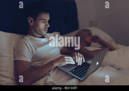 Sospettoso giovane uomo utilizzando computer portatile mentre ragazza dorme nel letto di notte Foto Stock