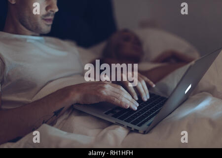 Ritagliato colpo di sospettoso giovane uomo utilizzando laptop mentre la moglie dorme nel letto di notte Foto Stock