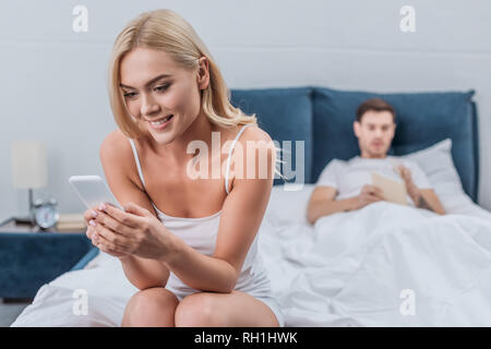 Sorridente giovane donna seduta sul letto e utilizza lo smartphone mentre fidanzato libro lettura dietro Foto Stock