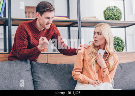 Iracondo del pugno di scuotimento e guardando spaventata ragazza tenendo lo smartphone a casa Foto Stock