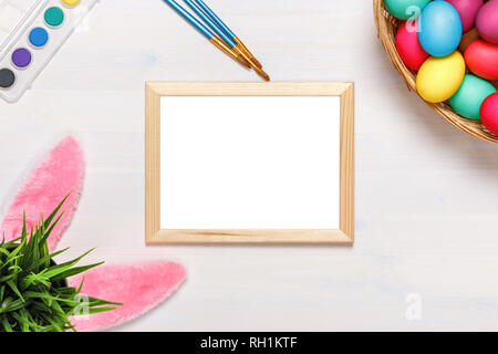 Un telaio, orecchie di coniglietto, un vaso di fiori artificiali con erba verde e colorati di uova in una scatola di cartone, vernici e pennelli. Concetto di pasqua. Spazio di copia Foto Stock