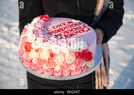 Bella torta con fiori di colore rosa nelle mani di una ragazza su uno sfondo di neve. La scritta sulla torta, tradotto dal russo: Buon compleanno, Foto Stock