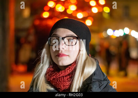 Giovane e bella ragazza in bicchieri di notte in posa contro lo sfondo di notte le luci. La bionda in un cappello nero è sempre sorridente. Foto Stock