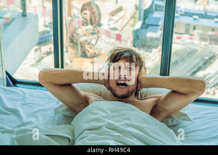 Giovane uomo disteso su un letto coperto le orecchie a causa del rumore. Nella finestra dopo il letto è possibile vedere la costruzione di una nuova casa Foto Stock