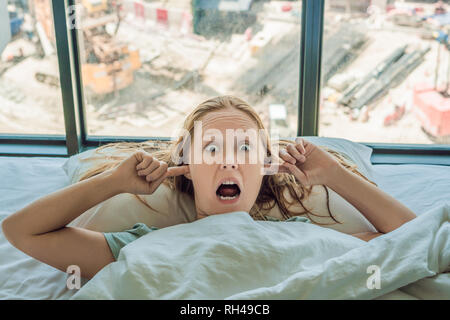 Giovane donna sdraiata su un letto coperto le orecchie a causa del rumore. Nella finestra dopo il letto è possibile vedere la costruzione di una nuova casa Foto Stock