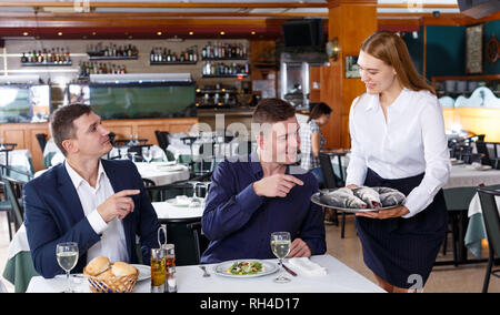 Cameriera in maglia bianca dimostrando il vassoio di pesce a due maschi i visitatori al ristorante Foto Stock