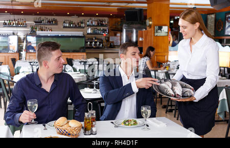 Cameriera in maglia bianca dimostrando il vassoio di pesce a due maschi i visitatori al ristorante Foto Stock