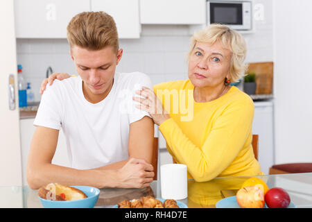 Senior donna abbracciando e sostenere moralmente il suo triste figlio adulto in cucina Foto Stock