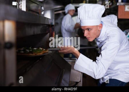 Ritratto di certi giovani chef lavorano nel moderno ristorante cucina, prepara la pizza fuori del forno Foto Stock
