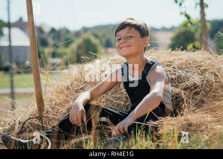 Ritratto fiducioso e sorridente ragazzo in tuta masticare fieno di sunny farm Foto Stock