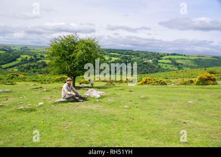 Giovani caucasici britannico escursionista seduto su un masso di granito da un albero di biancospino. Parco Nazionale di Dartmoor, Devon, Regno Unito. Foto Stock