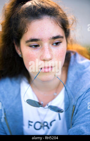 Un ritratto di un triste e pensieroso adolescente ragazza con le lentiggini e capelli lunghi marrone che detiene nella sua bocca un archetto di bicchieri e tristemente guardò verso il basso. Foto Stock