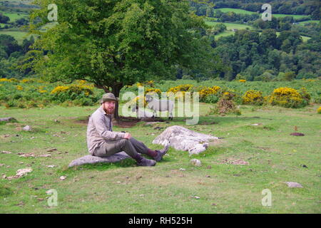 Giovani caucasici britannico escursionista seduto su un masso di granito da un albero di biancospino. Parco Nazionale di Dartmoor, Devon, Regno Unito. Foto Stock