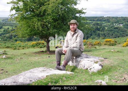 Giovani caucasici britannico escursionista seduto su un masso di granito da un albero di biancospino. Parco Nazionale di Dartmoor, Devon, Regno Unito. Foto Stock