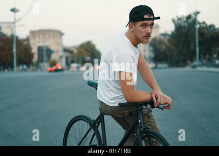Outdoor ritratto di un moderno e giovane uomo della strada, seduto sulla bici. Un giovane uomo di atletica che indossa i pantaloni khaki, T-shirt bianco, cercando di fotocamera. Foto Stock
