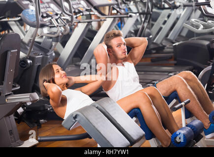 Ben addestrato giovane allenatore assistere allegro ragazza per fare esercizio di abs sit ups in palestra indoor . Focus sulla donna Foto Stock