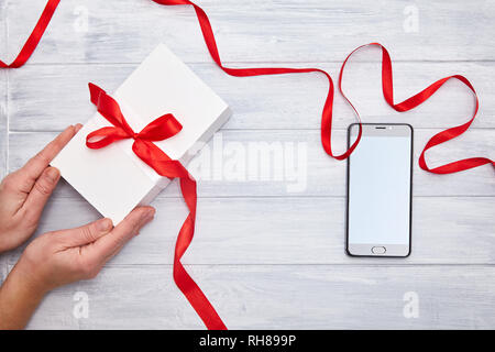 Scatola regalo bianca in mani con un nastro rosso e lo smartphone su un sfondo woodem. Compleanno, Natale, il giorno di San Valentino idea regalo Foto Stock