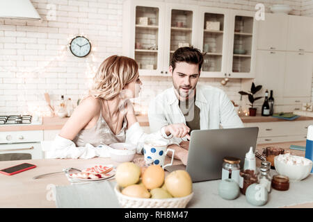 Barbuto bello uomo positivo in una camicia bianca che mostra le immagini su un computer portatile a sua moglie Foto Stock