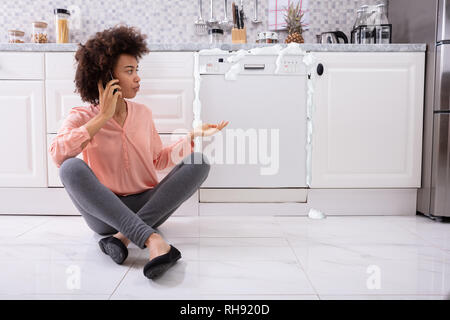 Triste donna tecnico di chiamata sul cellulare per fissare lavastoviglie con schiuma proveniente da esso Foto Stock