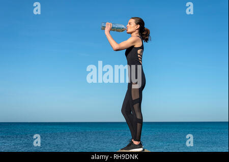 Sportive permanente sulla sommità di una roccia sul mare di bere da una bottiglia di plastica di acqua Foto Stock