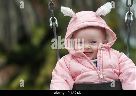 Close-up di 6 mese-vecchio baby ragazza sorridente su uno swing durante il periodo invernale presso la Del Rey Lagoon Park in Playa Del Rey, CA. Foto Stock