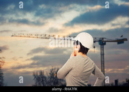 Vista posteriore di pensosa giovane donna ingegnere indossare casco protettivo tenendo la mano sotto il mento alla ricerca attenta alla gru a torre in constructio Foto Stock