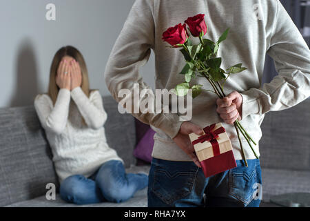 Mans mani nascondendo holding elegante bouquet di rose rosse e dono con nastro bianco dietro la schiena e la donna con le mani sul viso della sorpresa vi aspetta a letto Foto Stock