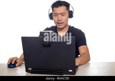 Maschio asiatico che indossano le cuffie la riproduzione di un video e il gioco online in streaming con una webcam su un pc portatile. L'immagine illustra industria di intrattenimento ed el Foto Stock