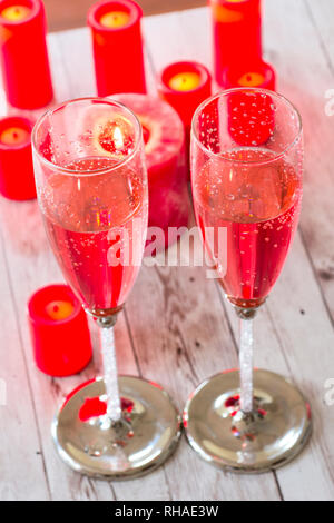 Due flauti champagne riempito con il colore rosa spumeggiante bevanda, sul piano portapaziente circondato con la masterizzazione di candele rosse. Foto Stock