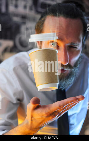 Far levitare in aria il bicchiere di carta con caffè caldo e di evaporazione da esso. Barista, un barbuto Giovane Uomo in camicia bianca con una cravatta, crea miracoli - Foto Stock