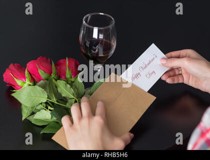 Ragazza mani tiene un messaggio felice il giorno di San Valentino sulla carta dal retro busta sul banco nero con rosso di bouquet di rose e vino rosso in vetro Foto Stock