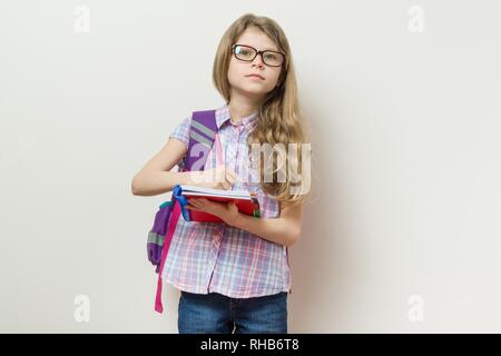 Giovani schoolgirl in bicchieri e con uno zaino scrive in una scuola di notebook. Sfondo parete luminosa nella scuola Foto Stock