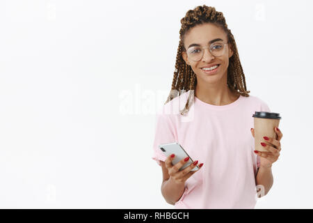 Ottenere eccitato con sorso di caffè. Amichevole-cercando americano africano femmina con cool timori azienda bicchiere di carta e lo smartphone sorridente Foto Stock