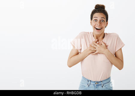 Grati e riconoscenti bella felice giovane donna con bun disordinati in maglietta e jeans tenendo le mani sul petto sorridente largamente con senza parole Foto Stock