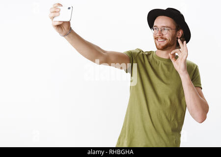 Studio shot di simpatici viaggiatori maschio in vacanza prendendo selfies sullo smartphone extanding mano con telefono mobile e sorridente alla schermata che mostra bene gesto Foto Stock